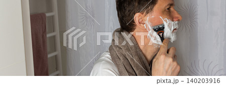 A 53 year old man shaves in the bathroom during his morning routine. He uses a razor and shaving cream while wearing a towel around his neck. Water is visible in the sink, banner A 53 year old man shaves in the bathroom during his morning routine. He uses a razor and shaving cream while wearing a towel around his neck. Water is visible in the sink, banner 140203916