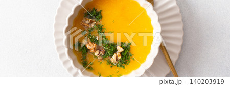 A bowl of pumpkin cream soup sits on a plate. The soup contains nuts and herbs. A yellow checkered napkin lies nearby. This meal can be enjoyed at breakfast or lunch, banner A bowl of pumpkin cream soup sits on a plate. The soup contains nuts and herbs. A yellow checkered napkin lies nearby. This meal can be enjoyed at breakfast or lunch, banner 140203919