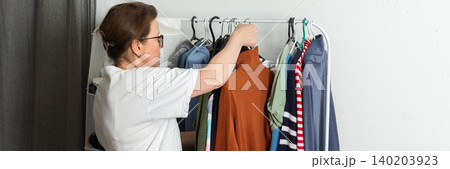 A 52-year-old woman sorts and hangs washed clothes on a hanger inside her home. She is organizing different garments after completing her laundry tasks, banner A 52-year-old woman sorts and hangs washed clothes on a hanger inside her home. She is organizing different garments after completing her laundry tasks, banner 140203923