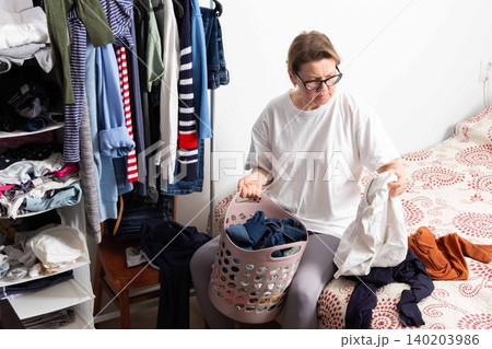 A person sits on the floor, sorting dirty clothes into a basket. The scene shows a cluttered laundry space with clothes piled on a chair and bed, indicating laundry day A person sits on the floor, sorting dirty clothes into a basket. The scene shows a cluttered laundry space with clothes piled on a chair and bed, indicating laundry day 140203986