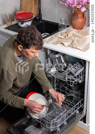 A man is putting dirty dishes into the dishwasher in his kitchen. He focuses on the task while several items rest in the appliance, ready for cleaning 140203988