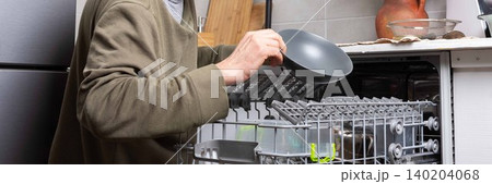 A man stands in his kitchen taking clean dishes out of the dishwasher. He focuses on the process of unloading the clean dishes and organizing them for use, banner 140204068