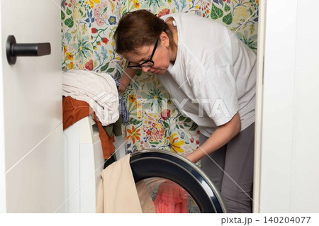 Woman stands in bathroom putting clothes into washing machine while surrounded by towels and laundry, carrying out daily household task with focus and care 140204077