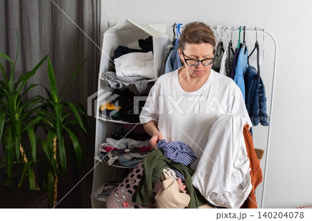 A person stands in a room holding a basket full of dirty clothes. Garments hang in the background. The setting is bright and casual, focusing on the laundry task ahead 140204078