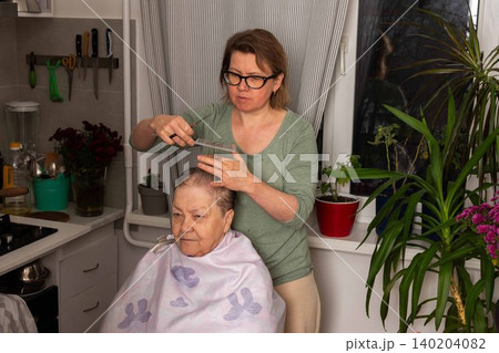 A woman uses a comb to cut her mother hair in the kitchen. They share a special moment together. The room is bright with plants nearby and tools hung on the wall A woman uses a comb to cut her mother hair in the kitchen. They share a special moment together. The room is bright with plants nearby and tools hung on the wall 140204082