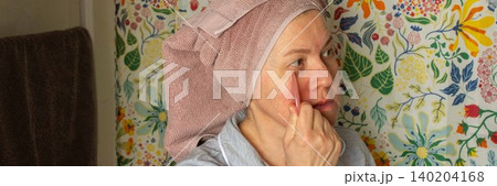 A 50-year-old woman follows her morning routine in the bathroom. She applies skincare products on her face while wearing a towel on her head and comfortable clothing, banner A 50-year-old woman follows her morning routine in the bathroom. She applies skincare products on her face while wearing a towel on her head and comfortable clothing, banner 140204168