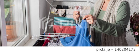 A woman around 50 years old is hanging clothes on a drying rack outside her balcony. She carefully organizes items to let them dry in the sunlight during the afternoon, banner A woman around 50 years old is hanging clothes on a drying rack outside her balcony. She carefully organizes items to let them dry in the sunlight during the afternoon, banner 140204175