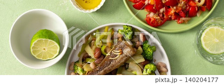 Breakfast includes baked hake placed on a bed of boiled beans with broccoli and peppers, accompanied by water and tomato salad on the table, banner Breakfast includes baked hake placed on a bed of boiled beans with broccoli and peppers, accompanied by water and tomato salad on the table, banner 140204177