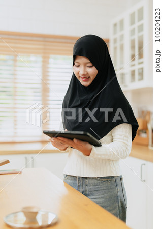 Young Arab Muslim woman in hijab works with tablet computer at home kitchen happy smiling. 140204223