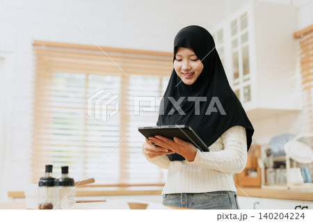 Young Arab Muslim woman in hijab works with tablet computer at home kitchen happy smiling. Young Arab Muslim woman in hijab works with tablet computer at home kitchen happy smiling. 140204224