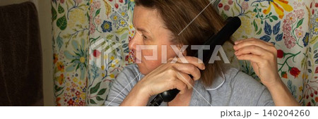 A 50-year-old woman styles her hair with a straightener in a bathroom. Floral curtains add color to the space. She is focused on her hair, preparing for the day ahead, banner 140204260