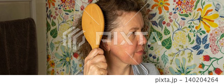 A 50-year-old woman stands in her bathroom holding a wooden hairbrush. She focuses on brushing her hair while facing a floral patterned wall, preparing for her day ahead, banner A 50-year-old woman stands in her bathroom holding a wooden hairbrush. She focuses on brushing her hair while facing a floral patterned wall, preparing for her day ahead, banner 140204264