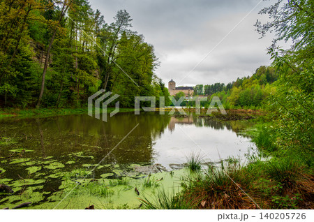 Kost Castle rises above a pond in the Bohemian Paradise region of the Czech Republic. Forests surround the landscape under cloudy skies, creating a scenic historic view. 140205726