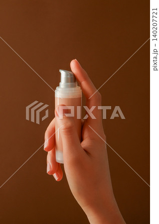 A hand holds a skincare bottle in front of a brown wall A hand holds a skincare bottle in front of a brown wall 140207721