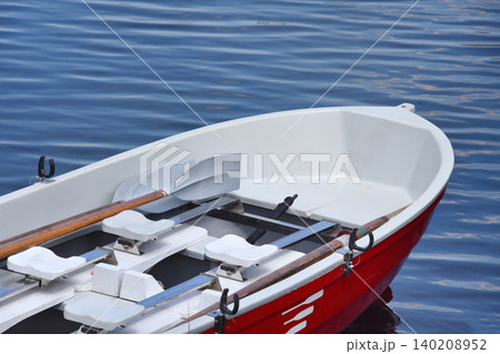 Red rowboat with wooden oars floats on calm blue water at a lakeside. Leisure craft with seats and oars suggests summer holidays and peaceful relaxation. Red rowboat with wooden oars floats on calm blue water at a lakeside. Leisure craft with seats and oars suggests summer holidays and peaceful relaxation. 140208952