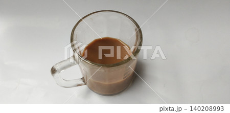 A simple close-up photo of a transparent glass cup containing a small amount of milk tea or coffee with milk, placed on a clean white background. A simple close-up photo of a transparent glass cup containing a small amount of milk tea or coffee with milk, placed on a clean white background. 140208993