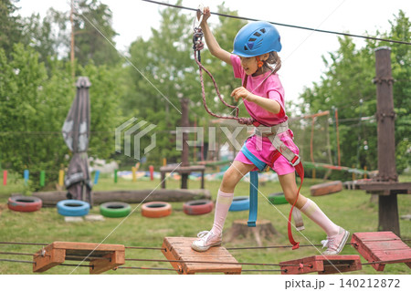 A child steps on the steps of a suspension bridge in a rope entertainment sports town. The girl holds on to the safety rope, moving the carbine A child steps on the steps of a suspension bridge in a rope entertainment sports town. The girl holds on to the safety rope, moving the carbine 140212872