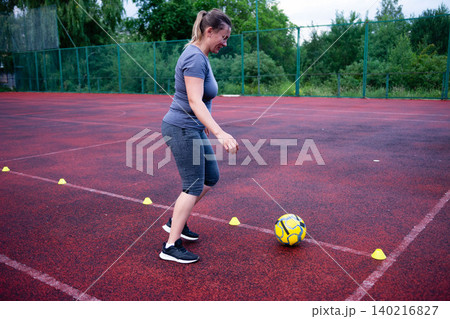 Woman practicing soccer drills on a red court. She dribbles the ball around cones, focusing on agility and ball control. 140216827