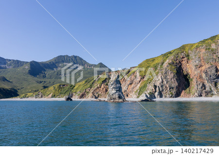 Rocky sea cliffs and green mountains on Kamchatka coast under clear blue sky Rocky sea cliffs and green mountains on Kamchatka coast under clear blue sky 140217429