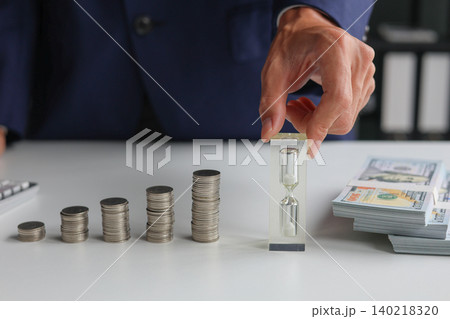 The concept of time and money. Hand holding a small hourglass next to stacks of coins and cash. The concept of time and money. Hand holding a small hourglass next to stacks of coins and cash. 140218320