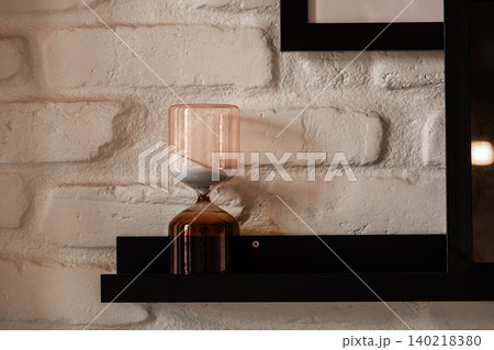 Modern Glass Hourglass on Black Shelf Against White Brick Wall 140218380