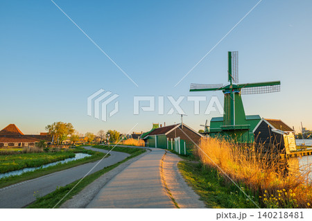 Scenic dutch landscape with traditional green windmill in zaanse schans at sunset, winding countryside road along water canal, historic wooden houses and famous holland tourism landmark Scenic dutch landscape with traditional green windmill in zaanse schans at sunset, winding countryside road along water canal, historic wooden houses and famous holland tourism landmark 140218481