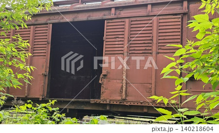 Old rusty red freight train car with open door surrounded by fresh green leaves on foreground. Atmosphere of abandoned industrial transportation Old rusty red freight train car with open door surrounded by fresh green leaves on foreground. Atmosphere of abandoned industrial transportation 140218604