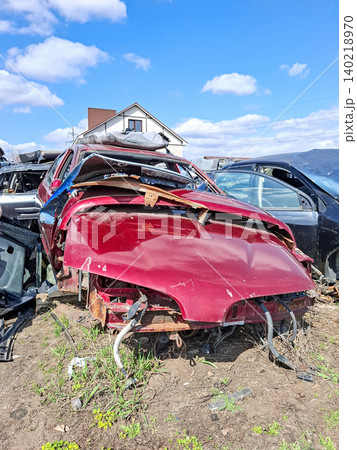 A crimson car body sits in a junkyard with a smashed hood. The car is among other scrap vehicles under a clear sky. This scene represents the aftermath of a traffic accident and recycling efforts. 140218970