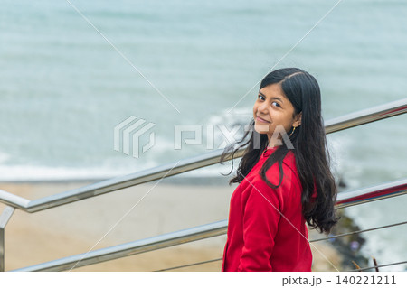 A teen in a red jacket stands on a railing overlooking the ocean 140221211