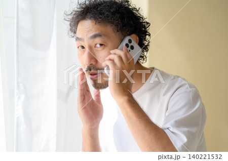 窓際でスマートフォンで小声で通話する日本人男性 窓際でスマートフォンで小声で通話する日本人男性 140221532