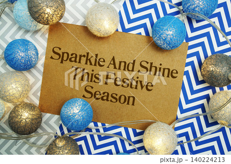 Sparkle And Shine This Festive Season text written on paper card with LED cotton balls decorations on blue background Sparkle And Shine This Festive Season text written on paper card with LED cotton balls decorations on blue background 140224213