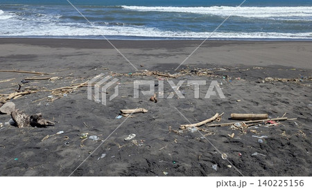 Scattered driftwood, plastic waste, organic debris litter a dark volcanic sand beach. Foaming white waves roll onto the shore from the blue ocean. This image highlights the environmental impact of Scattered driftwood, plastic waste, organic debris litter a dark volcanic sand beach. Foaming white waves roll onto the shore from the blue ocean. This image highlights the environmental impact of 140225156