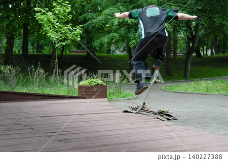 An unrecognizable young man balancing on his skateboard during a trick on the edge of a wooden ramp. Full-length back view shot with lush green trees and park pathway in the background on a summer 140227388