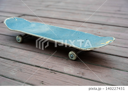 A close-up shot of an old blue skateboard with scratches on a dark wooden board surface. The camera angle is low, showcasing the wheels, trucks, and the texture of the deck. 140227431