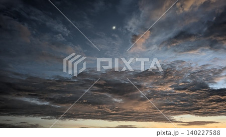 Wide scenic view captures heavy textured dark altocumulus cloud layers at dawn. A small crescent moon shines in blue morning skies, creating a beautiful dramatic natural background template. 140227588