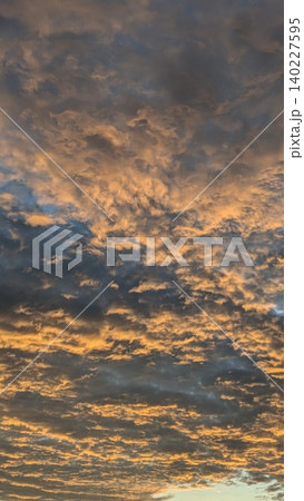 Vertical view captures heavy textured altocumulus cloud layers at sunrise. Golden orange light illuminates dramatic dark moody sky patterns, creating a beautiful natural backdrop layout template. Vertical view captures heavy textured altocumulus cloud layers at sunrise. Golden orange light illuminates dramatic dark moody sky patterns, creating a beautiful natural backdrop layout template. 140227595