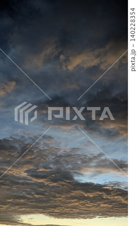 Vertical scenic view captures heavy textured dark storm cloud layers shifting at dawn. Soft golden sunrise light warms the lower horizon, creating a beautiful dramatic natural backdrop template. 140228354
