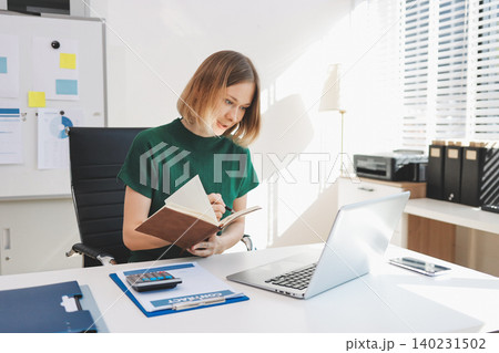 Concept of business working, Businesswoman sitting while writing and checking business analytic document  report  and holding tablet with computer pc, smartphone on desk in her workstation. 140231502