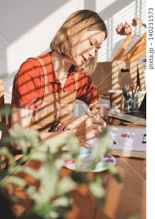Picture of serious concentrated young Caucasian female artist sitting at desk with painting accessories, holding tube of oil paint, mixing colors on palette; unfinished painting on canvas near her Picture of serious concentrated young Caucasian female artist sitting at desk with painting accessories, holding tube of oil paint, mixing colors on palette; unfinished painting on canvas near her 140231573