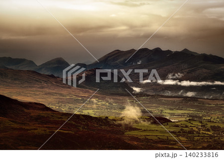 Sunlight streams through dark clouds, illuminating parts of a valley and casting shadows on distant mountain ranges. 140233816