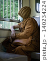 Wrapped in the soft, textured folds of traditional saffron robes, a Buddhist monk sits quietly by a train window, his gaze fixed on the world passing outside the barred frame. Wearing a knit beanie 140234224