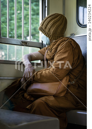 Wrapped in the soft, textured folds of traditional saffron robes, a Buddhist monk sits quietly by a train window, his gaze fixed on the world passing outside the barred frame. Wearing a knit beanie Wrapped in the soft, textured folds of traditional saffron robes, a Buddhist monk sits quietly by a train window, his gaze fixed on the world passing outside the barred frame. Wearing a knit beanie 140234224