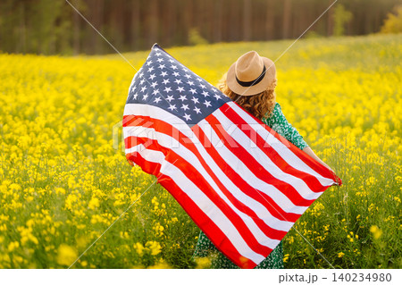 Beautiful girl in hat with American flag in sunflower field. Freedom. Sunset light The girl smiles. Beautiful girl in hat with American flag in sunflower field. Freedom. Sunset light The girl smiles. 140234980