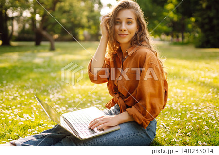 Woman in casual clothes with a laptop sits on lawn on sunny day. Happy freelancer working outdoors. Woman in casual clothes with a laptop sits on lawn on sunny day. Happy freelancer working outdoors. 140235014