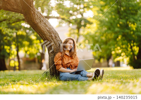 Woman in casual clothes with a laptop sits on lawn on sunny day. Happy freelancer working outdoors. 140235015