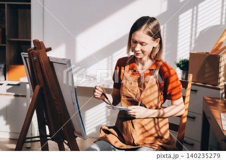 Picture of serious concentrated young Caucasian female artist sitting at desk with painting accessories, holding tube of oil paint, mixing colors on palette; unfinished painting on canvas near her Picture of serious concentrated young Caucasian female artist sitting at desk with painting accessories, holding tube of oil paint, mixing colors on palette; unfinished painting on canvas near her 140235279