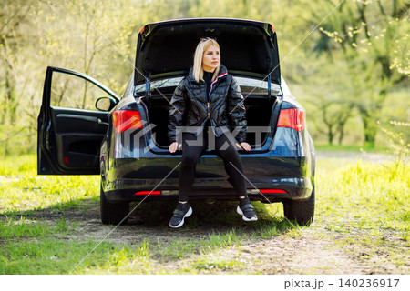 fair haired woman in a black jacket sits in the open trunk of a blue automobile in a clearing fair haired woman in a black jacket sits in the open trunk of a blue automobile in a clearing 140236917