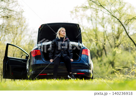 woman in dark clothing sits in the open trunk of a blue car against the background of trees from below woman in dark clothing sits in the open trunk of a blue car against the background of trees from below 140236918