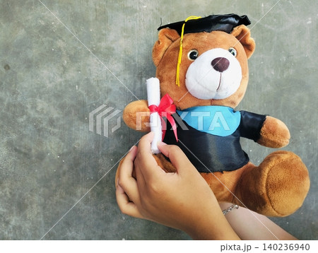 Hand Holding Graduation Teddy Bear with Diploma on Concrete Background Hand Holding Graduation Teddy Bear with Diploma on Concrete Background 140236940