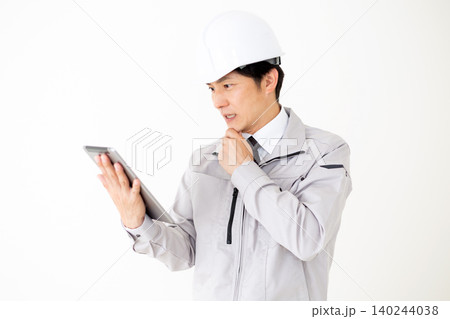 タブレットを見て考えるヘルメットをかぶったグレーの作業着のミドル男性ビジネスマン白バック 140244038